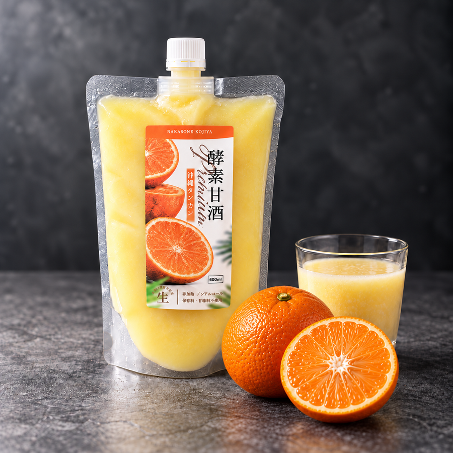 黒い石の台に置いた酵素甘酒 Premium 沖縄タンカンのパウチと、グラスに注いだ淡い黄色の甘酒、丸ごとと半分に切ったタンカン